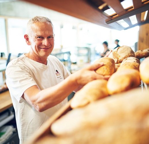 KI generiert: Ein Mann im Bäcker-Outfit präsentiert verschiedene Brote in einer Bäckerei.