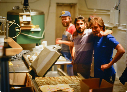 KI generiert: Drei Männer in einer Fabrikumgebung, lachend und posierend an einem Arbeitstisch mit verpackten Produkten.