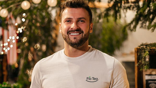 KI generiert: Ein lächelnder Mann in einem weißen "Fio Tea" T-Shirt vor unscharfem Hintergrund mit Lichtern und Pflanzen.